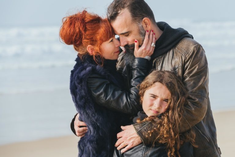 Une femme aux cheveux roux et au manteau de fourrure bleu marine touche doucement le visage de sa sœur barbue vêtue d'une veste en cuir marron alors qu'ils s'embrassent sur une plage. Une jeune fille aux cheveux bruns bouclés se tient devant l'homme, portant également une veste sombre, avec l'océan et le ciel en arrière-plan.