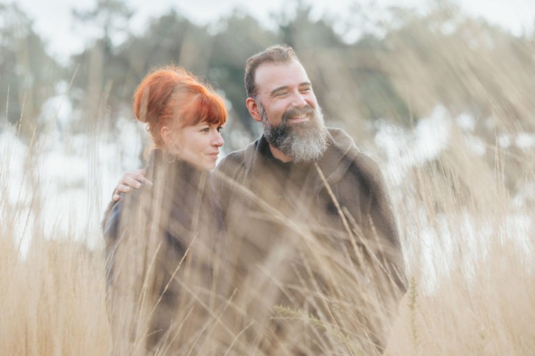 Scène extérieure mettant en scène un couple souriant au milieu d'herbes hautes et sèches. La femme, aux cheveux roux vifs, se penche vers l'homme, qui a une barbe fournie et des cheveux gris. Tous deux portent des vêtements sombres. L'arrière-plan est constitué d'arbres flous et d'un ciel nuageux, créant une atmosphère sereine et douce pour ces âmes sœurs.