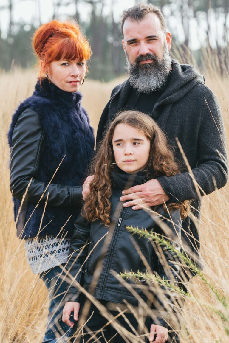 Une famille de trois personnes, véritables âmes sœurs, se tient debout dans un champ d'herbe haute et sèche. La femme a les cheveux roux attachés et porte un gilet de fourrure sombre sur un haut à manches longues. L'homme a une barbe fournie, des cheveux noirs et porte une veste sombre. L'enfant aux cheveux longs et bouclés et à la veste noire se tient devant eux. Des arbres sont visibles à l'arrière-plan