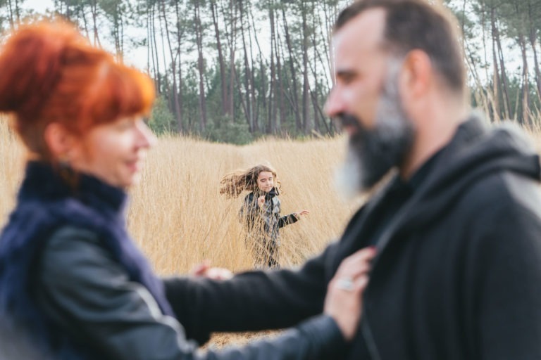 Au premier plan, un couple se tient par la main, flou, et se regarde comme deux âmes sœurs. À l'arrière-plan, une jeune fille aux cheveux longs marche dans l'herbe haute et sèche, le regard tourné vers l'objectif. Des arbres sont visibles au loin. L'image présente un décor serein et extérieur.