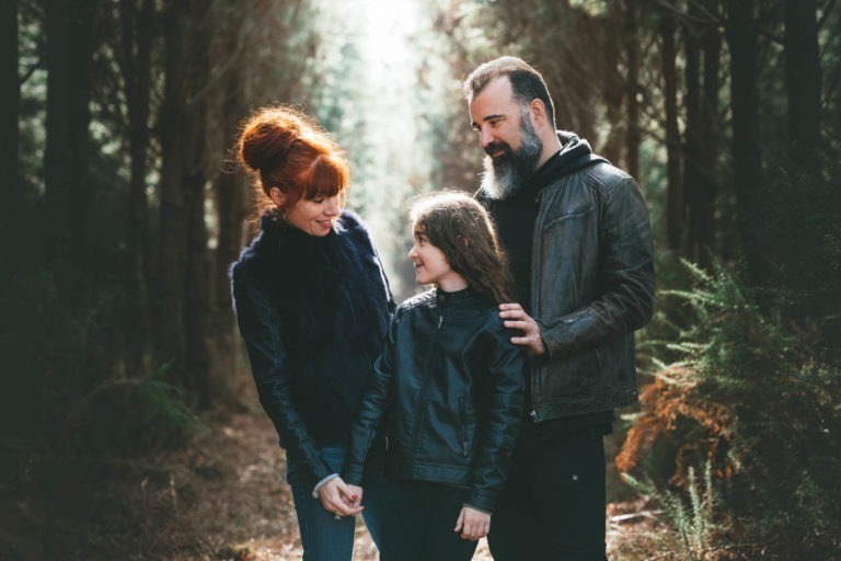 Une famille de trois personnes se tient dans une clairière, éclairée par la lumière du soleil filtrant à travers les arbres. Chaque membre de la famille porte une tenue sombre et coordonnée. Les parents tiennent doucement la main de leur enfant, tous trois se souriant chaleureusement comme des âmes sœurs. La scène est sereine et intime, entourée par la nature.