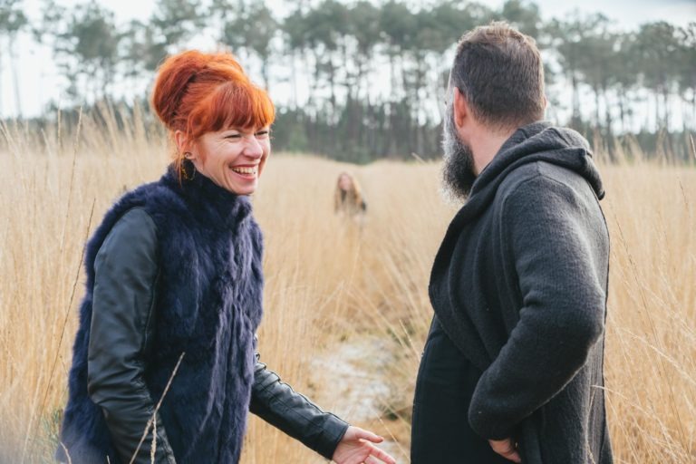 Une femme aux cheveux roux, vêtue d'un gilet en fourrure bleue, rit et tend la main vers un homme barbu portant un sweat-shirt à capuche gris. Debout dans un champ d'herbe haute et sèche avec des arbres à feuilles persistantes au loin, leur connexion joyeuse suggère qu'ils sont âmes sœurs. Une silhouette floue s'attarde à l'arrière-plan.