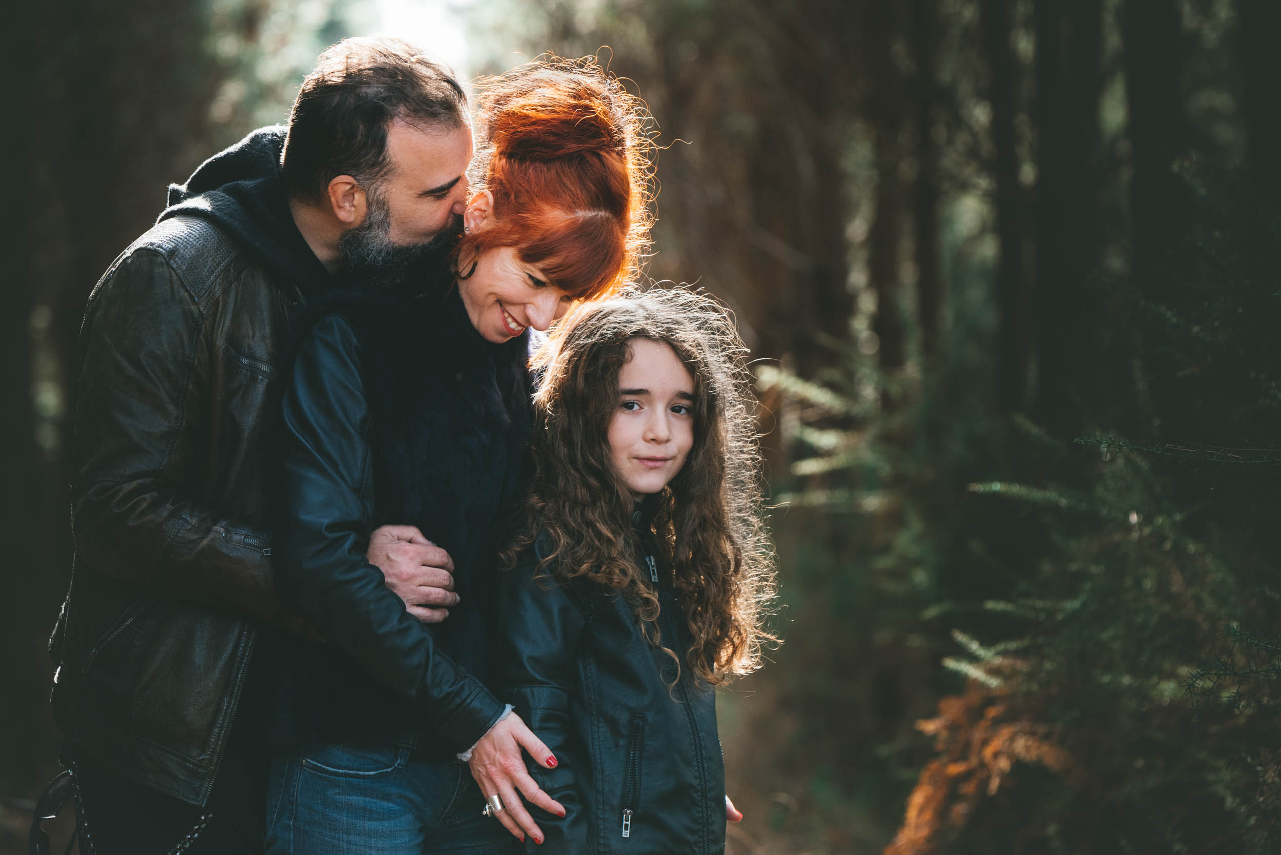 Un homme barbu embrasse une femme aux cheveux roux sur la tête tout en la tenant contre lui. Ils portent tous les deux des vestes en cuir, comme de véritables âmes sœurs. Une jeune fille aux cheveux bouclés se tient devant eux, regardant la caméra. Ils sont dans une forêt où la lumière du soleil filtre à travers les arbres.