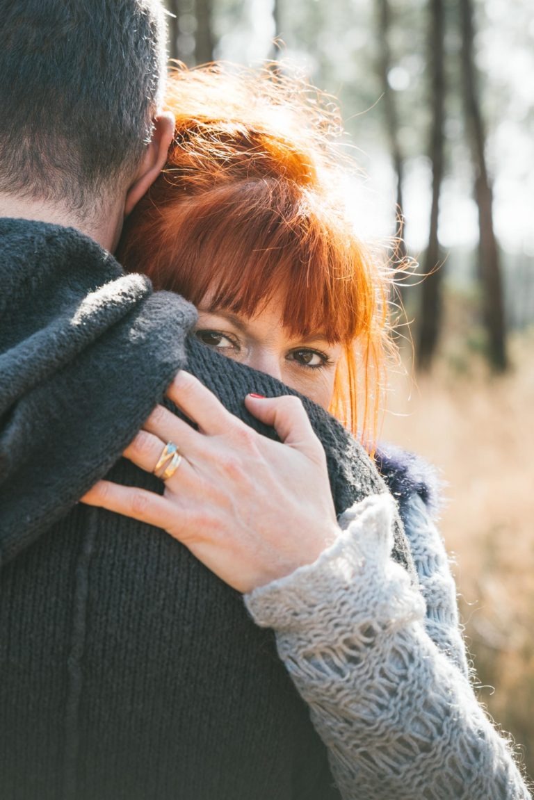 Une femme aux cheveux roux enlace un homme tout en regardant directement l'objectif, capturant un moment de tendresse entre âmes sœurs. Elle porte un pull en tricot et une bague à la main gauche, qui repose sur le dos de l'homme. L'arrière-plan est flou, montrant un décor extérieur avec de grands arbres et la lumière du soleil filtrant à travers.