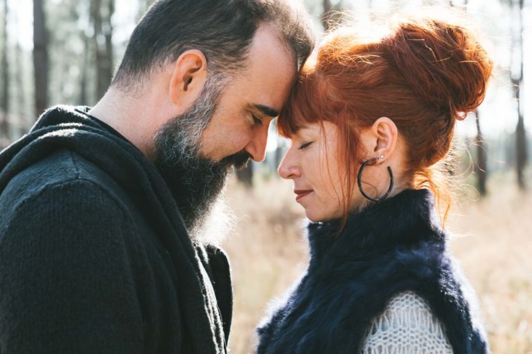 Un couple se tient debout, le front contre le mur, les yeux fermés, dans un décor extérieur serein. L'homme, avec une barbe fournie et un pull foncé, et la femme, portant un gilet bleu en fourrure sur un pull clair, avec ses longs cheveux roux coiffés en chignon lâche. Comme des âmes sœurs, entourées d'arbres flous et de soleil.