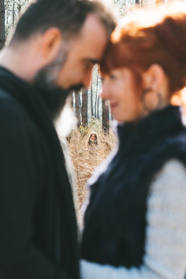 Un homme et une femme, âmes sœurs, se tiennent côte à côte, face à face, leurs fronts se touchant dans une zone boisée. Tous deux sont légèrement flous. À l'arrière-plan, une jeune fille aux cheveux longs jette un œil derrière les hautes herbes, souriant au couple tandis que la lumière du soleil filtre à travers les arbres.