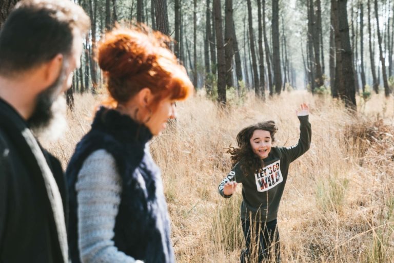 Un enfant court joyeusement dans une forêt ensoleillée, parsemée de grands arbres fins et d'herbe sèche. Au premier plan, une femme aux cheveux roux et un homme barbu, qui apparaissent comme des âmes sœurs, se tiennent à la lisière de la forêt et observent l'enfant. L'enfant porte un pull gris avec l'inscription « Let's Go Wild » et a les cheveux longs et foncés.