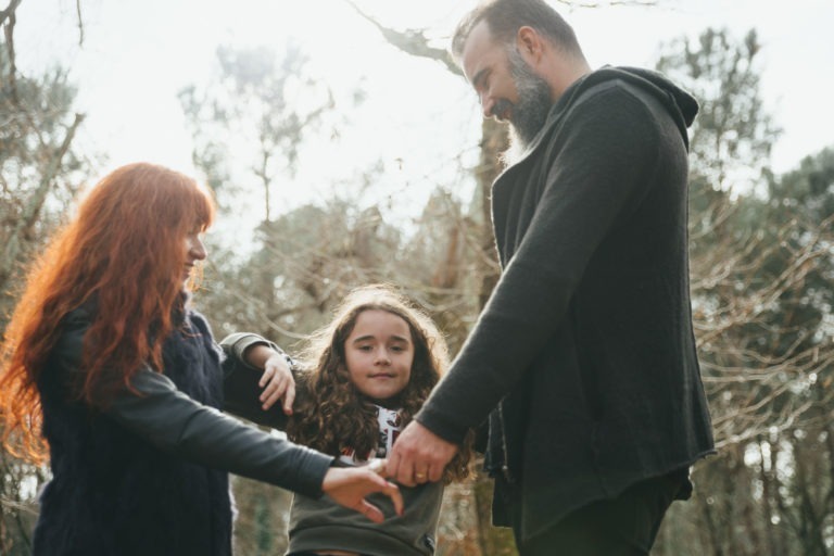 Une famille de trois personnes se tient la main à l'extérieur. La femme à gauche a de longs cheveux roux, tandis que l'homme à droite a une barbe. Au centre se trouve un jeune enfant aux cheveux longs et bouclés. Ils sourient tous et semblent apprécier la compagnie des autres comme des âmes sœurs sous les arbres baignés de soleil.