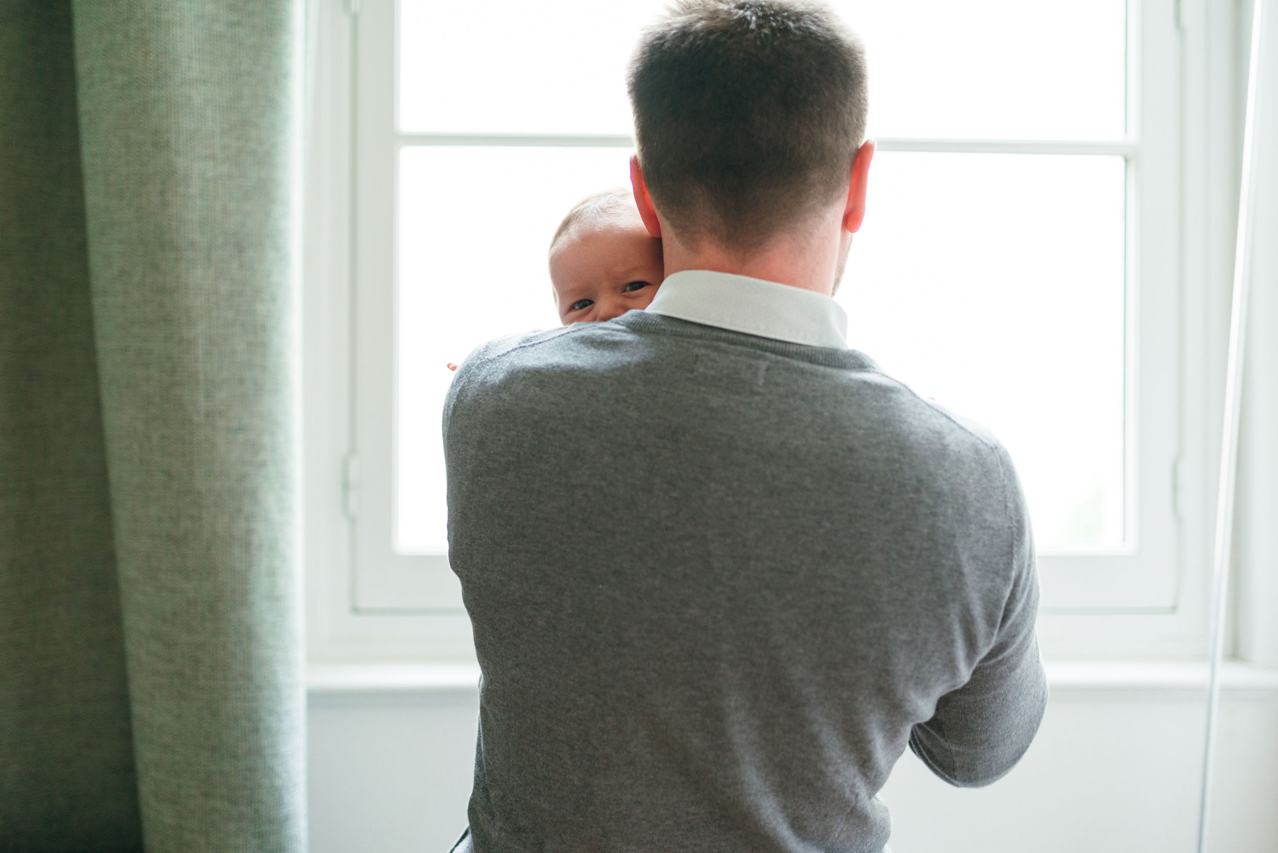 Un homme aux cheveux bruns courts, vêtu d'un pull gris et d'une chemise blanche à col, se tient dos à la caméra, tenant doucement un bébé sur son épaule gauche. Le bébé, qui regarde par-dessus l'épaule de l'homme avec de grands yeux et un léger sourire, crée une explosion de sensations devant une fenêtre à cadre blanc avec des rideaux lumineux.