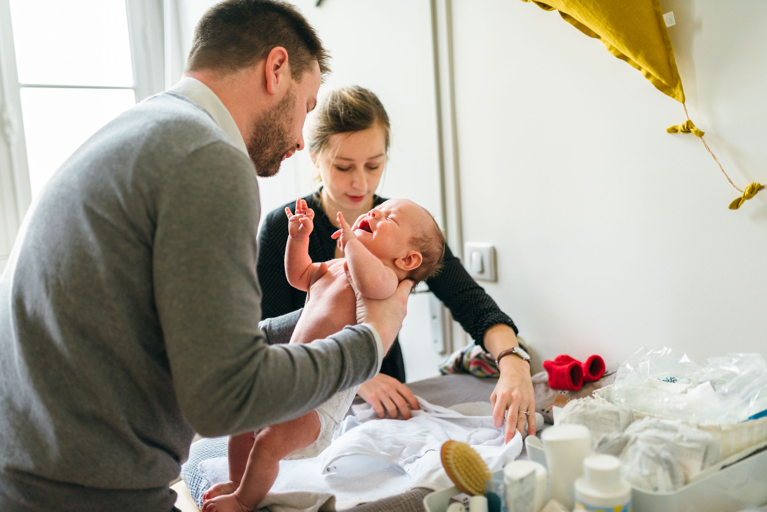 Un homme et une femme changent la couche d'un bébé sur une table à langer. L'homme tient doucement le bébé, tandis que la femme prépare des articles sur la table remplie de couches, de lingettes, de lotions et de vêtements. La lumière naturelle provenant d'une fenêtre voisine illumine la scène, créant une explosion de sensations de chaleur et d'attention.