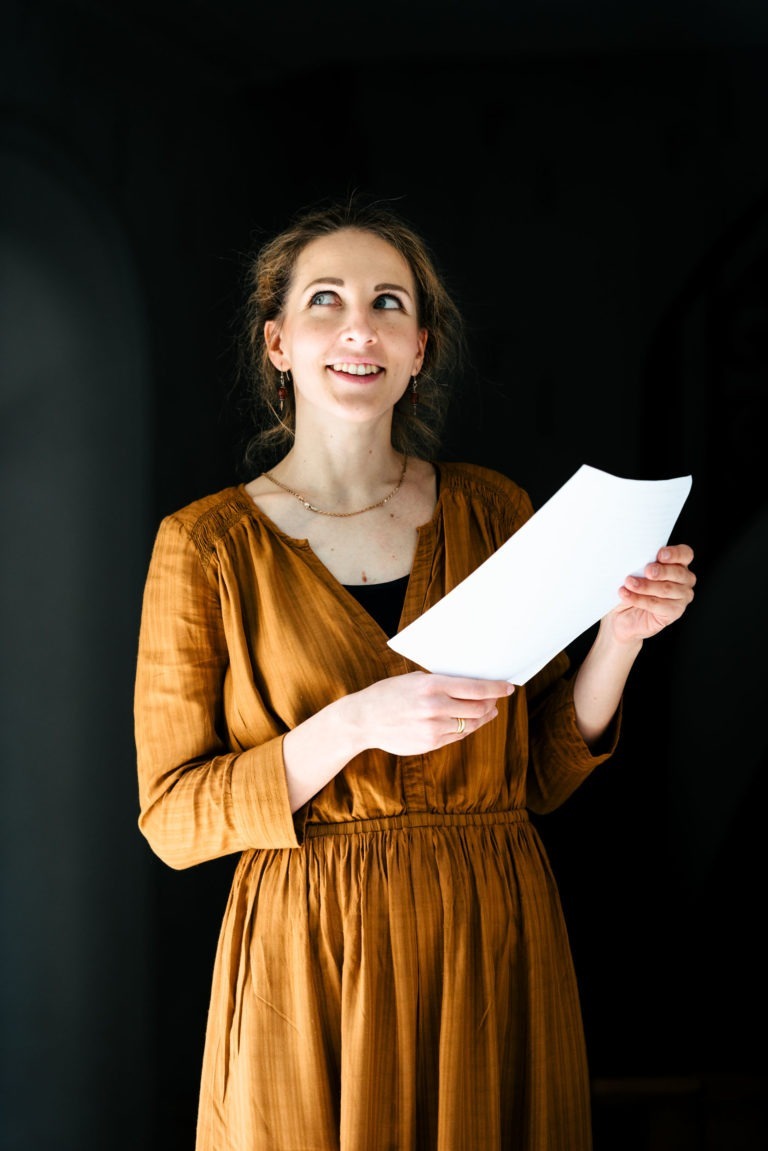 Une femme aux cheveux châtain clair, vêtue d'une robe à manches longues couleur moutarde, se tient debout sur un fond sombre. Elle regarde vers le haut et légèrement vers la droite tout en tenant une feuille de papier blanc dans sa main gauche. L'expression de son visage semble pensive ou contemplative, rappelant une performance du duo Altalina en Gironde.