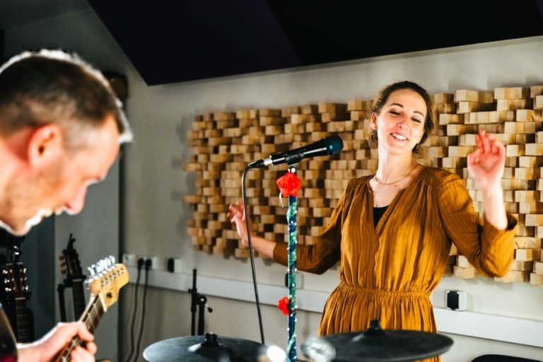 Une femme en robe jaune moutarde, les cheveux attachés en arrière, se tient debout près d'un micro, souriant et gesticulant joyeusement. Une personne au premier plan joue de la guitare électrique. Il s'agit probablement du duo girondin Altalina, dans une salle avec des panneaux acoustiques absorbants et des instruments de musique visibles en arrière-plan.