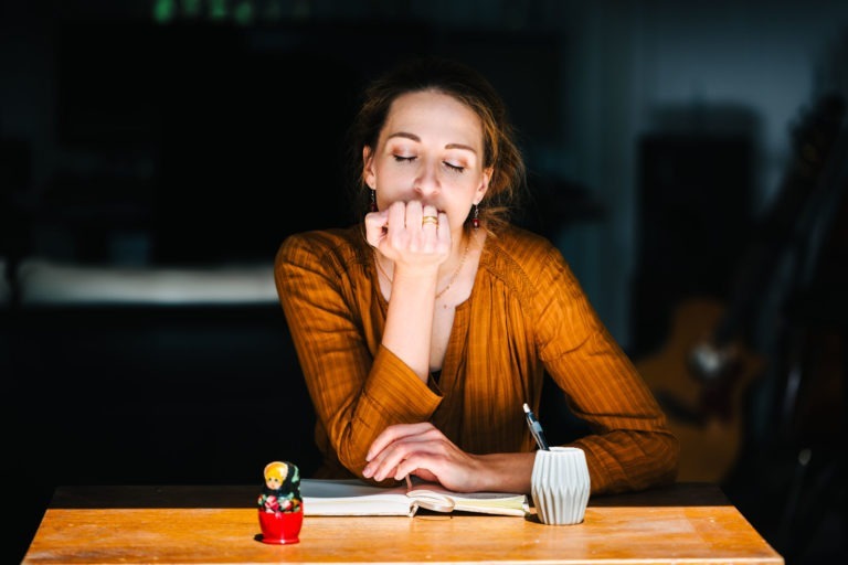 Une femme, le visage posé sur sa main, est assise à une table faiblement éclairée, un livre fermé devant elle. Elle porte un chemisier orange et a les yeux fermés, semblant contemplative ou fatiguée. Sur la table se trouvent une petite poupée décorative et un porte-stylo avec un stylo à l'intérieur. À l'arrière-plan, une guitare musicale du groupe Altalina est partiellement visible.