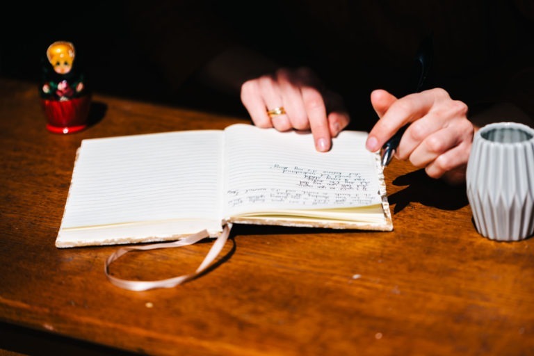 Un cahier ouvert aux pages lignées est posé sur une table en bois. Une personne, dont on ne voit que les mains, écrit dans le cahier avec un stylo. Près du cahier se trouvent une petite poupée russe et une tasse en céramique grise à nervures de la Gironde. Un marque-page en ruban pend au bord du cahier.