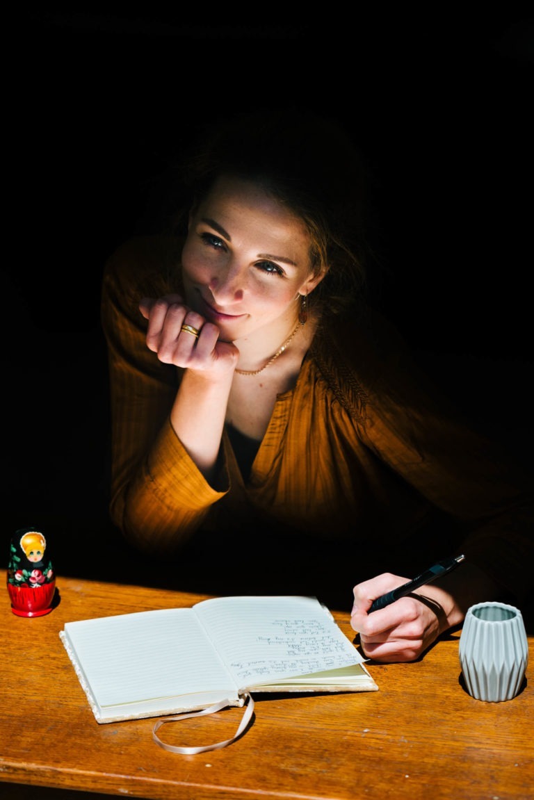 Une personne aux cheveux bruns s'appuie sur sa main tout en écrivant dans un cahier ouvert sur une table en bois. Elle porte un haut couleur moutarde et est éclairée par un projecteur. Sur la table se trouvent une petite poupée Matriochka colorée et un vase décoratif blanc. Une mélodie subtile de la comédie musicale du duo Altalina remplit le fond sombre.