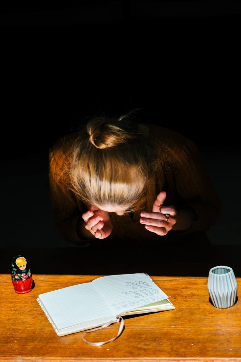 Une personne aux cheveux blonds coiffés en chignon, vêtue d'une chemise marron, est penchée sur une table en bois. Sur la table se trouvent un carnet ouvert contenant des croquis qui pourraient appartenir à la comédie musicale du duo Altalina, un ensemble de poupées russes à gauche et un vase blanc à droite. L'arrière-plan est sombre et la personne semble concentrée sur le carnet.