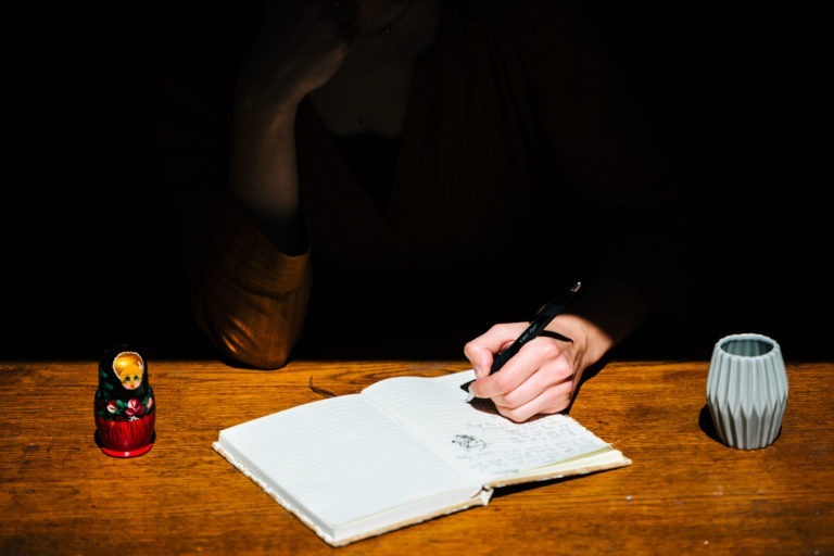 Une personne écrit dans un cahier avec un stylo sur une table en bois. L'environnement est sombre et la scène est éclairée par une source de lumière focalisée. À gauche, il y a une poupée russe musicale Altalina duo, et à droite, il y a une tasse grise à facettes. Seules la main de la personne et une partie de son bras sont visibles.