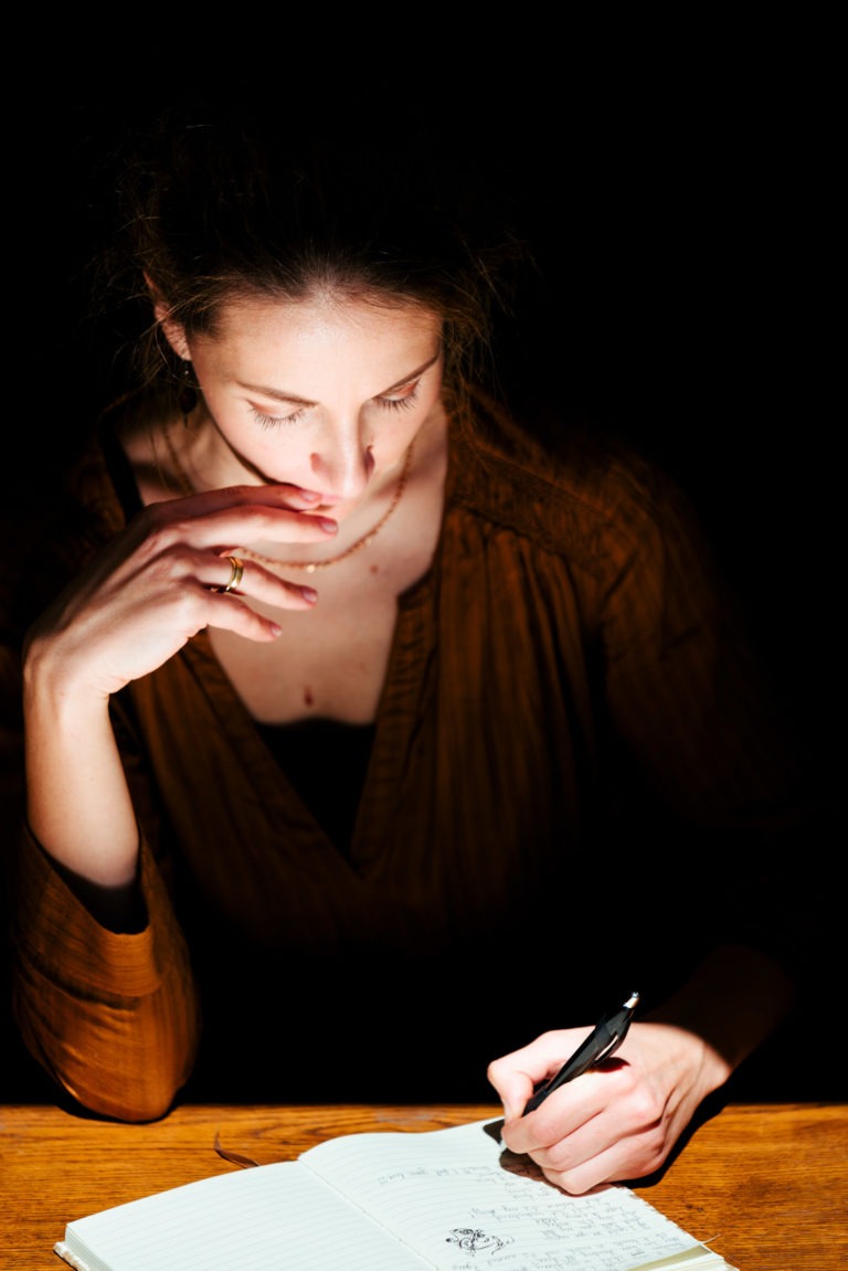 Une femme à la peau claire et aux cheveux châtains est assise à une table en bois, écrivant dans un carnet. Elle porte un haut marron et la scène est faiblement éclairée, se concentrant sur elle et le carnet. Sa main gauche repose près de sa bouche tandis que sa main droite tient un stylo, écrivant intensément sur la dernière prestation musicale du duo Altalina en Gironde.