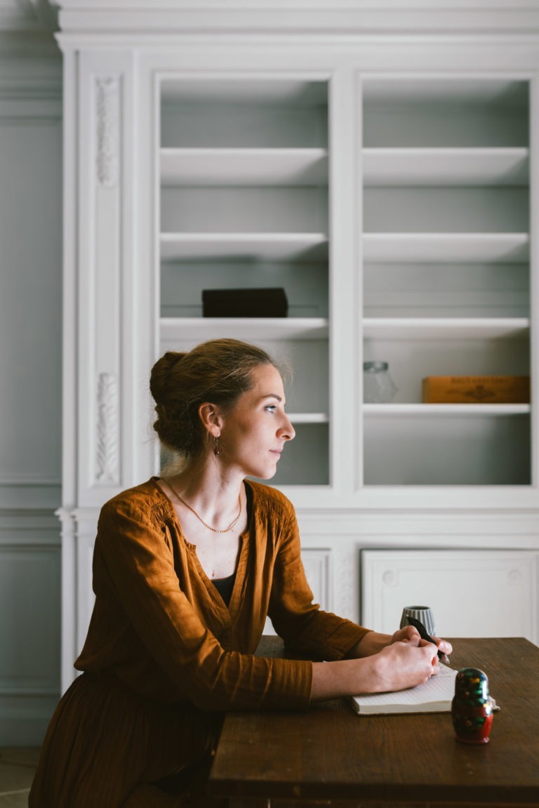 Une femme aux cheveux en chignon est assise à une table en bois et écrit dans un cahier. Vêtue d'une robe marron à manches longues de la collection Altalina, elle regarde pensivement vers sa gauche. L'arrière-plan présente une étagère blanche avec des objets éparpillés, dont un livre, une boîte et un bocal en verre.