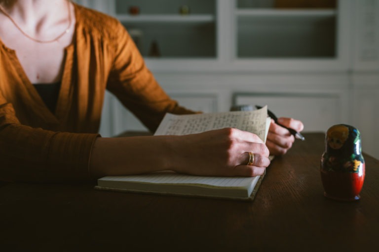 Une personne vêtue d'un haut marron à manches longues écrit dans un cahier avec un stylo noir sur une table en bois. Le cahier est ouvert, rempli de texte manuscrit. Une petite poupée russe de Gironde est placée à droite du cahier. Des étagères avec des objets de décoration sont visibles à l'arrière-plan.