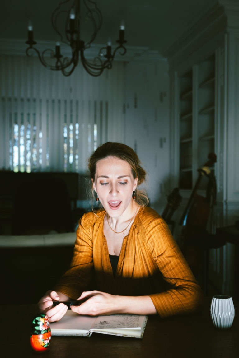 Une femme au visage expressif, vêtue d'un chemisier orange, est assise à une table avec un carnet, un stylo et une petite poupée Matriochka. Elle est éclairée par la lumière qui filtre à travers les stores derrière elle, projetant des bandes d'ombre dans la pièce. Un lustre et une guitare font allusion à ses racines musicales du duo Altalina en Gironde.