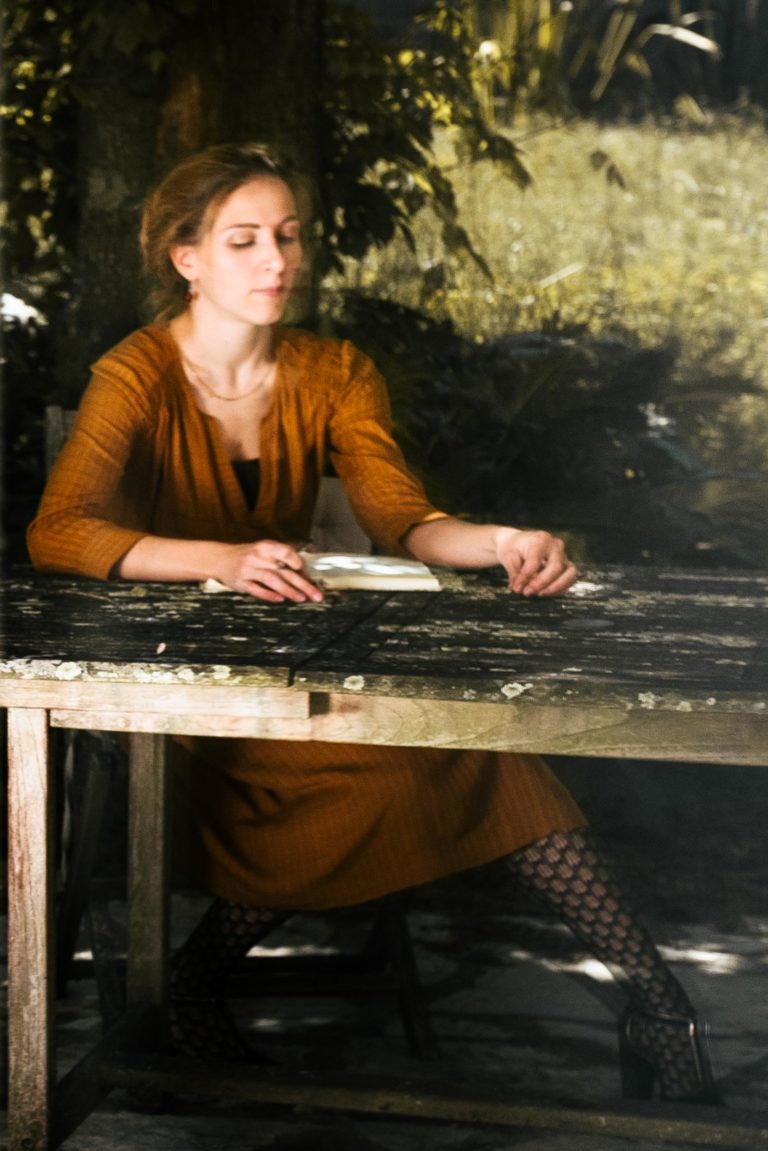 Une femme vêtue d'une robe orange brûlée à manches longues est assise à une table en bois rustique en plein air en Gironde. La table est patinée par les intempéries et elle est à l'ombre d'un arbre. Ses yeux sont fermés comme si elle écoutait une comédie musicale du duo Altalina, son bras droit reposant sur la table et ses collants à motifs visibles en dessous.