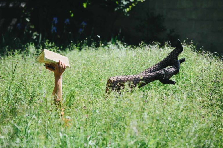 Une image montre une personne allongée dans l'herbe haute, seuls les bras et les jambes étant visibles. Une main tient un objet rectangulaire de couleur dorée, tandis que les jambes, chaussées de chaussures noires à talons hauts et de bas résille, pointent vers le haut. Dans la verdure luxuriante de la Gironde, la scène évoque une pochette d'album pour un groupe musical du groupe Altalina.