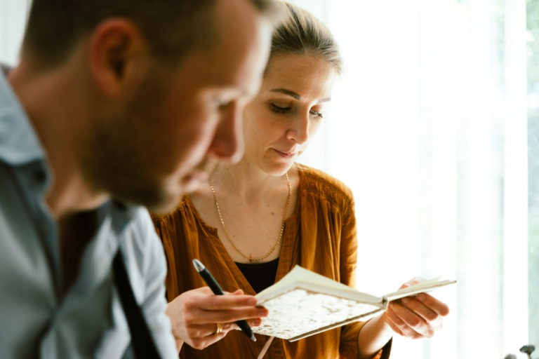 Un homme et une femme sont assis à l'intérieur, la femme étant au centre de l'attention. Elle tient un carnet ouvert dans sa main gauche et un stylo dans sa main droite, semblant prendre des notes. Vêtue d'un chemisier marron, elle semble profondément impliquée. L'arrière-plan doucement éclairé crée une atmosphère intime proche d'une séance musicale du duo Altalina.