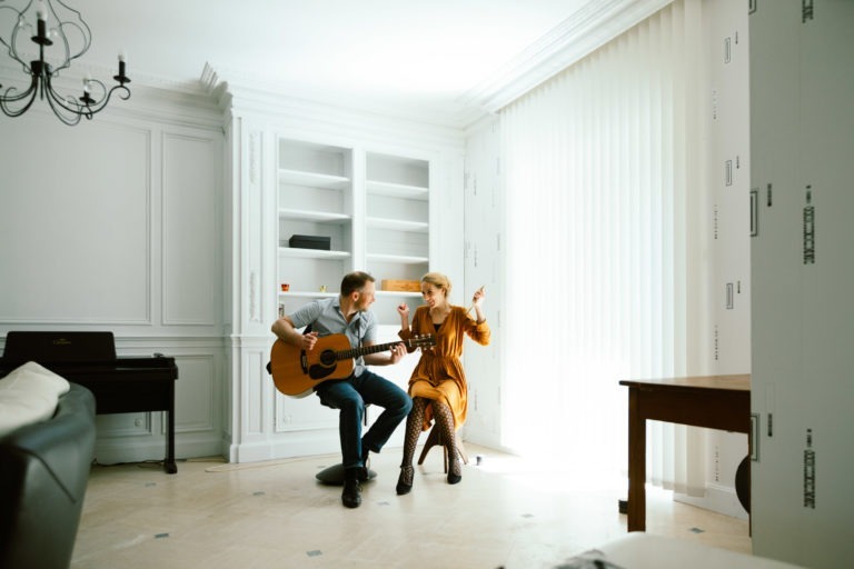 Un homme et une femme, connus sous le nom de duo Altalina, sont assis et jouent de la guitare dans une pièce lumineuse aux murs et aux meubles blancs. L'homme gratte la guitare tandis que la femme chante ou fait des gestes avec ses mains. Cette scène musicale se déroule dans une pièce dotée d'un piano, d'un canapé, d'une table, d'étagères et de grands stores verticaux couvrant une fenêtre.