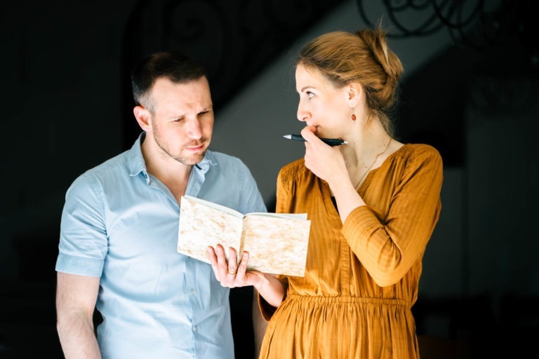 Un homme en chemise bleu clair et une femme en robe moutarde se tiennent debout côte à côte. La femme, l'air pensif, tient un carnet ouvert et un stylo près de sa bouche. L'homme regarde le carnet avec une expression sérieuse. Ils sont à l'intérieur avec un éclairage tamisé en arrière-plan, ressemblant au duo Altalina se préparant pour leur prochain concert en Gironde.