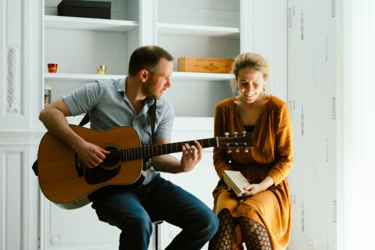 Un homme et une femme du duo Altalina sont assis à l'intérieur. L'homme, à gauche, joue de la guitare acoustique et regarde la femme, qui tient un livre et sourit. La femme porte une robe marron et des collants à motifs. Des étagères blanches et une caisse en bois sont visibles dans leur confortable maison girondine en arrière-plan.