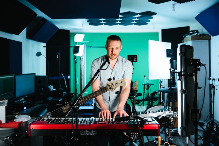 Un homme en chemise bleu clair joue du clavier et de la guitare électrique dans son home-studio, représentant le duo musical Altalina de Gironde. Il se tient devant un écran vert, entouré d'équipements tels que des panneaux d'éclairage, des moniteurs et des microphones. La pièce est remplie de divers équipements musicaux et d'enregistrement, notamment des batteries et des mixeurs audio.