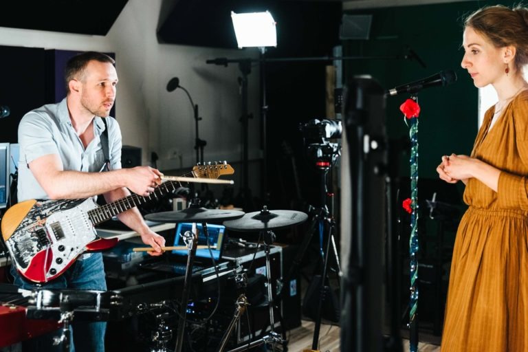 Un homme avec une guitare pointe une baguette vers une femme tenant un micro dans le studio de musique bien éclairé du duo Altalina. Divers équipements et instruments les entourent. La femme, vêtue d'une robe couleur moutarde, se tient debout près d'un pied de micro orné de fleurs rouges.