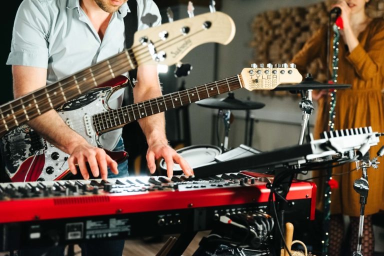 Une personne joue des deux mains sur un clavier rouge. Un autre individu gratte une guitare électrique avec enthousiasme. En arrière-plan, un chanteur en tenue couleur moutarde tient un micro. Une batterie et du matériel de studio sont également visibles, suggérant une séance musicale animée du duo Altalina en Gironde.