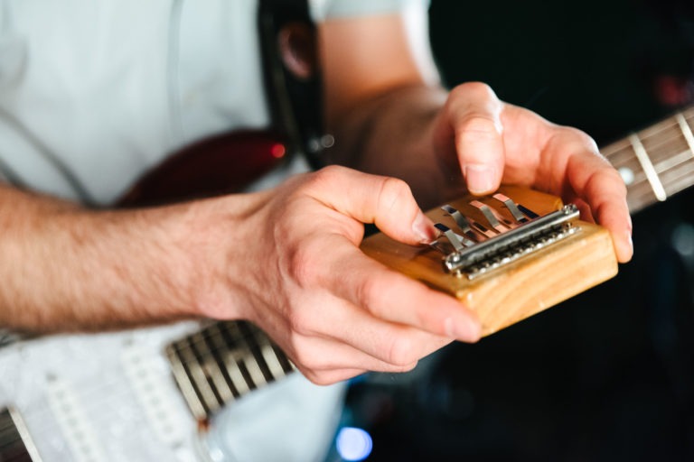 Gros plan de deux mains jouant du kalimba, un petit instrument de musique africain à lames métalliques montées sur un socle en bois. La personne qui tient le kalimba, qui fait partie de la comédie musicale du duo Altalina, porte également une sangle de guitare, ce qui indique qu'elle joue d'une guitare électrique partiellement visible en arrière-plan.