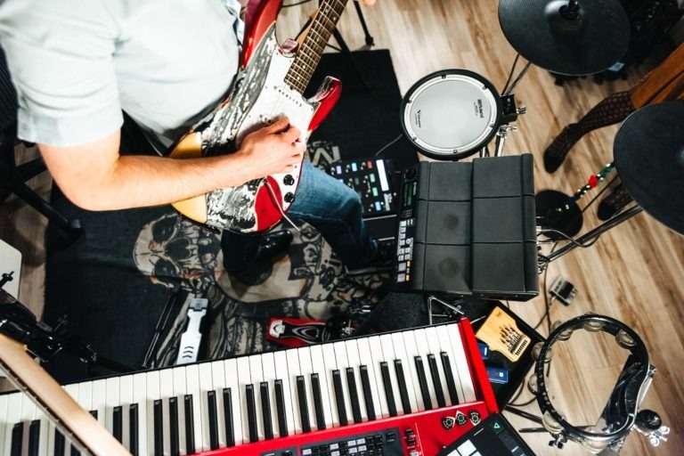 Une personne en chemise bleu clair joue de la guitare électrique rouge, debout près d'un clavier, de pads de batterie électroniques et d'autres équipements musicaux. Prise en perspective, la scène met en valeur le décor encombré du duo Altalina sur un plancher en bois en Gironde, mettant en valeur le grain du bois et un tapis.