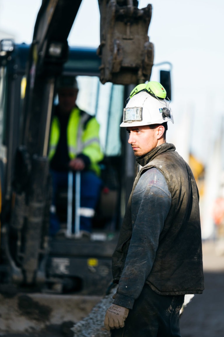 Un ouvrier du bâtiment portant un casque de sécurité blanc avec un cache-oreilles de sécurité vert et une veste EOVEST foncée se tient sur un chantier, regardant vers sa gauche. Derrière lui, il y a un ouvrier assis qui conduit une excavatrice. La scène semble se dérouler à l'extérieur en plein jour.