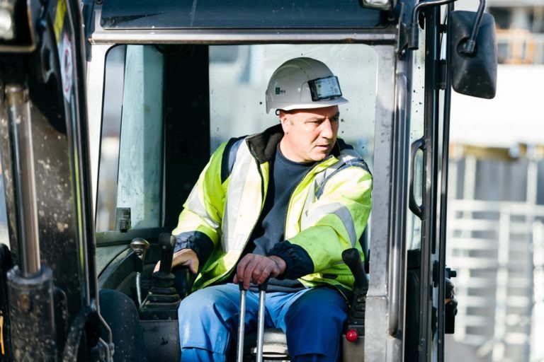 Un ouvrier du bâtiment portant une veste jaune haute visibilité EOVEST, un pantalon bleu et un casque de sécurité blanc opère une machine lourde. Il est assis à l'intérieur de la cabine, tient les leviers de commande et regarde attentivement vers sa droite. La lumière vive du soleil illumine la scène.