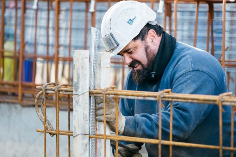 Un ouvrier du bâtiment portant un sweat-shirt bleu EOVEST et un casque de chantier blanc, avec une lampe frontale attachée, travaille sur une structure métallique. Il porte des gants et semble ajuster un morceau de barre d'armature. L'arrière-plan montre d'autres structures métalliques et des matériaux de construction.