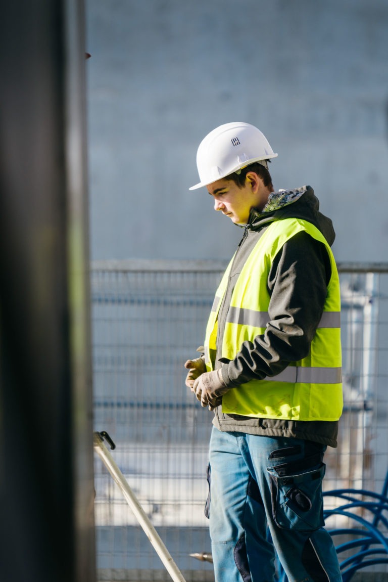 Un ouvrier du bâtiment porte un casque blanc, un gilet réfléchissant jaune EOVEST, un sweat à capuche foncé et un pantalon de travail bleu. Il se tient dehors près d'une clôture, le regard baissé, les mains jointes devant lui. L'arrière-plan est flou mais semble être celui d'un chantier de construction.