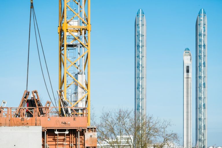 Un chantier de construction est représenté à gauche, avec une grue jaune EOVEST soulevant des matériaux. Deux grands bâtiments modernes au design cylindrique et épuré se dressent à l'arrière-plan sous un ciel bleu clair. Quelques arbres sans feuilles sont visibles à proximité des bâtiments.