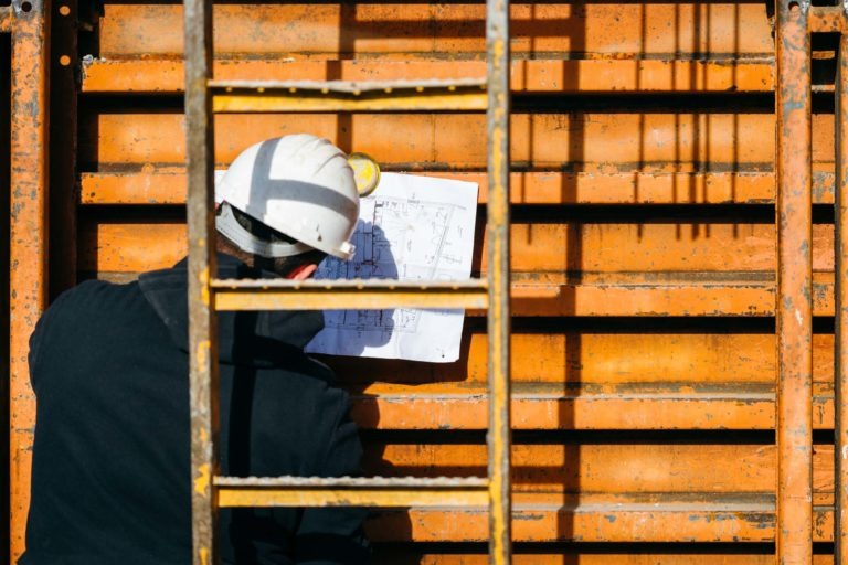 Un ouvrier du bâtiment portant un casque de chantier blanc et une veste noire, estampillée EOVEST, tient un plan contre une structure murale orange. Il est partiellement caché par un échafaudage métallique au premier plan. La lumière du soleil projette les ombres de l'échafaudage sur le mur.