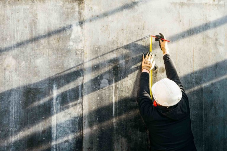 Un ouvrier du bâtiment portant un casque de sécurité EOVEST blanc et des vêtements noirs mesure un mur en béton à l'aide d'un mètre ruban et d'un marqueur rouge. L'ouvrier porte des gants de protection. Les ombres des poutres créent des lignes angulaires sur le mur, ajoutant un intérêt visuel à la scène.