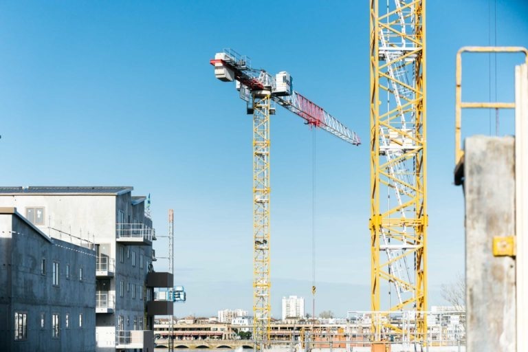 Un chantier de construction avec deux grandes grues jaunes, dont l'une est marquée EOVEST, dominant la zone. L'une des grues a une section horizontale rouge et blanche. Autour des grues se trouvent des bâtiments partiellement construits et des échafaudages. Le ciel est bleu clair et des bâtiments lointains sont visibles en arrière-plan.