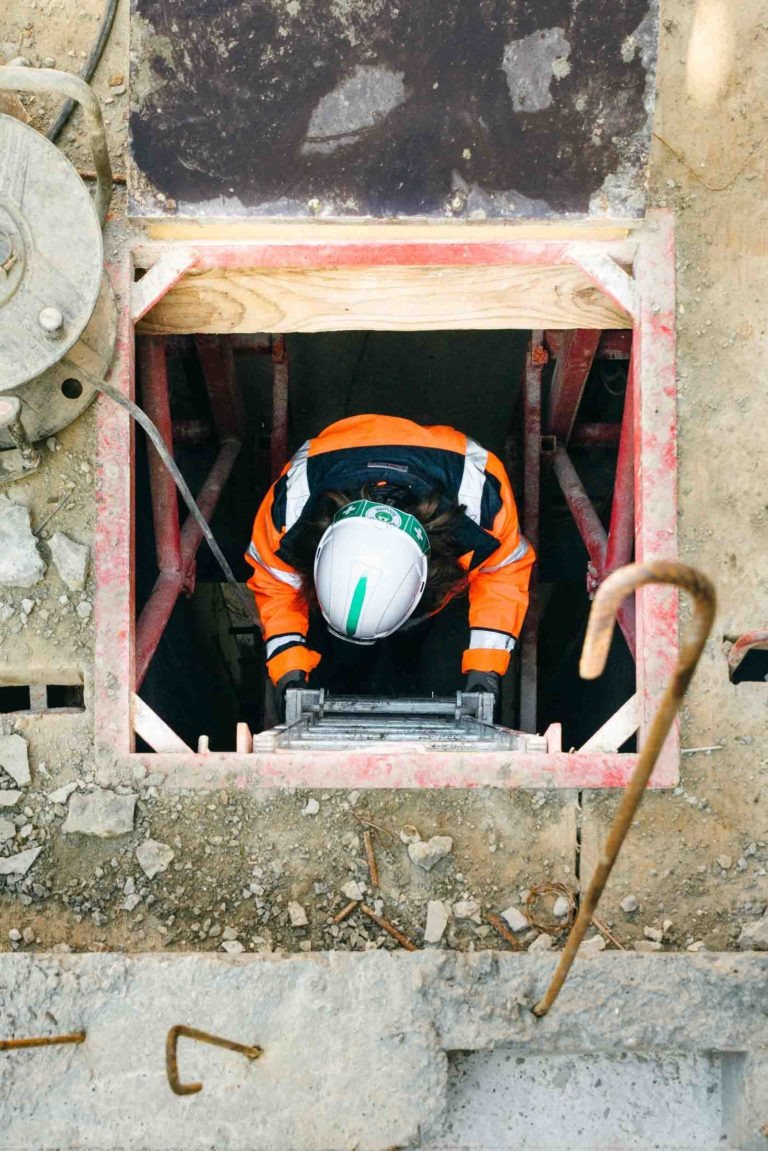 Un ouvrier du bâtiment en uniforme orange et noir EOVEST et casque de chantier blanc monte une échelle dans un puits carré entouré d'échafaudages. Le sol est poussiéreux et parsemé de matériaux de construction. L'ouvrier est vu d'en haut, le haut de son corps émergeant par l'ouverture.