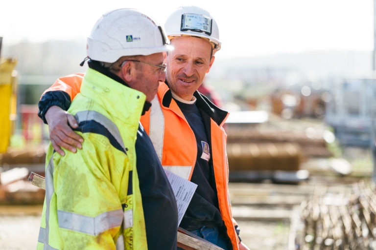 Deux ouvriers du bâtiment discutent sur un chantier. L'un, vêtu d'une veste jaune haute visibilité EOVEST, tient des documents. L'autre, vêtu d'une veste orange à bandes réfléchissantes, passe son bras autour du premier ouvrier en souriant. Tous deux portent des casques de sécurité blancs. L'arrière-plan est flouté : des équipements et des matériaux de construction.