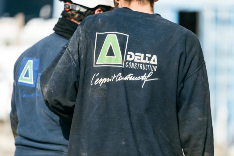 Deux ouvriers du bâtiment, vus de dos, portent des vestes EOVEST bleu marine avec le logo « Delta Construction ». Le logo comporte un triangle vert et gris à côté du texte « Delta Construction » avec le slogan « l'esprit bâtisseur ! » en dessous. Les vestes sont légèrement poussiéreuses.