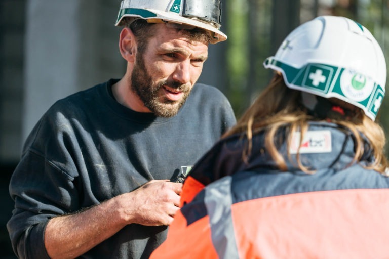 Un ouvrier du bâtiment barbu, vêtu d'une chemise noire et d'un casque de chantier blanc, discute avec une autre personne portant un casque de chantier blanc EOVEST avec des symboles verts et une veste orange haute visibilité. Tous deux sont sur un chantier de construction, avec des éléments d'arrière-plan flous.