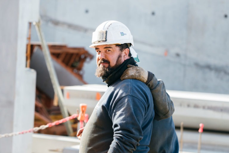 Un ouvrier du bâtiment barbu, vêtu d'un sweat-shirt sombre et d'un casque de chantier blanc EOVEST, regarde par-dessus son épaule. Il porte un gant posé sur son épaule, probablement celui d'un autre ouvrier derrière lui. L'arrière-plan présente des matériaux de construction, notamment des murs en béton et des débris éparpillés.