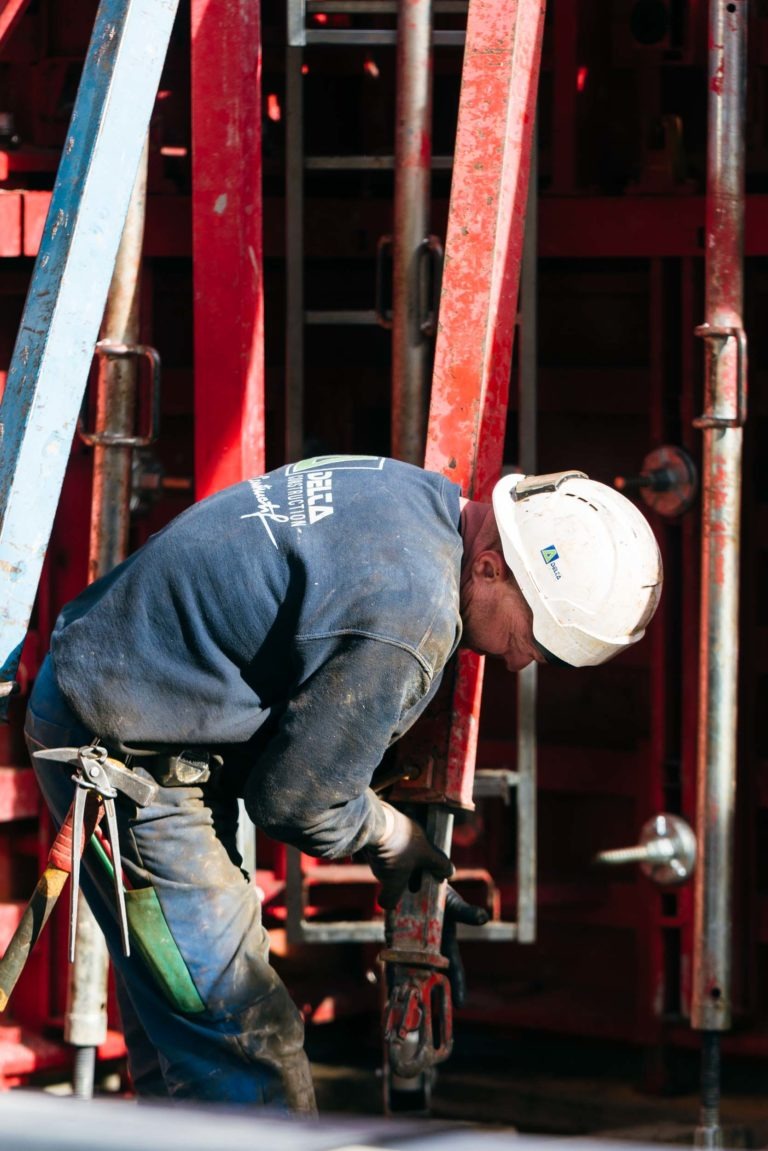 Un ouvrier du bâtiment portant un casque de sécurité blanc, une chemise à manches longues bleue et un harnais EOVEST est vu en train de travailler sur un échafaudage en métal rouge. L'ouvrier est penché, concentré sur le réglage d'un équipement avec ses mains. L'arrière-plan est rempli d'échafaudages et de matériaux de construction supplémentaires.